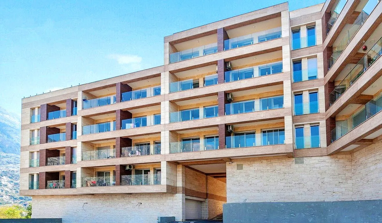 Exterior view of a multi-story residential building featuring balconies with glass railings against a mountain backdrop.