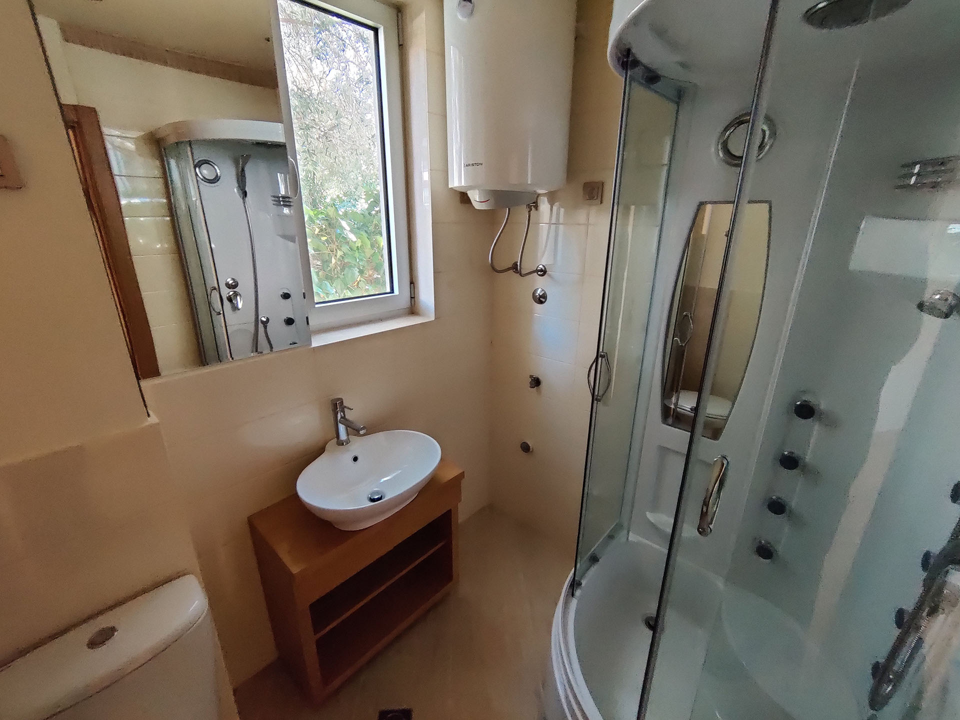 Bathroom in an apartment in Becici, Montenegro: shower cabin, vanity sink, water heater, and a window overlooking greenery.
