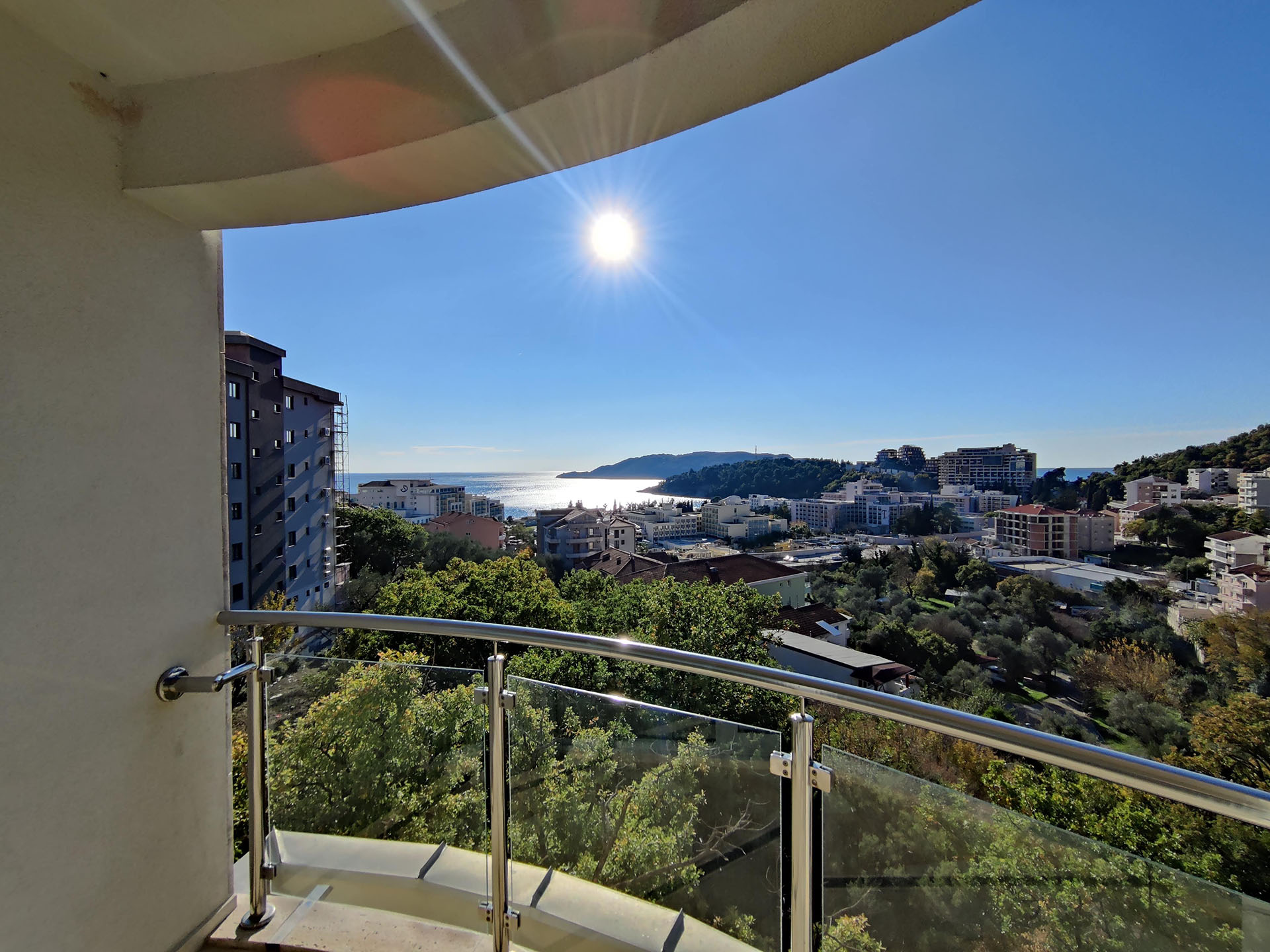 View from the apartment balcony in Bečići, Montenegro: a panorama of the city, green hills, and the Adriatic Sea on a sunny day.