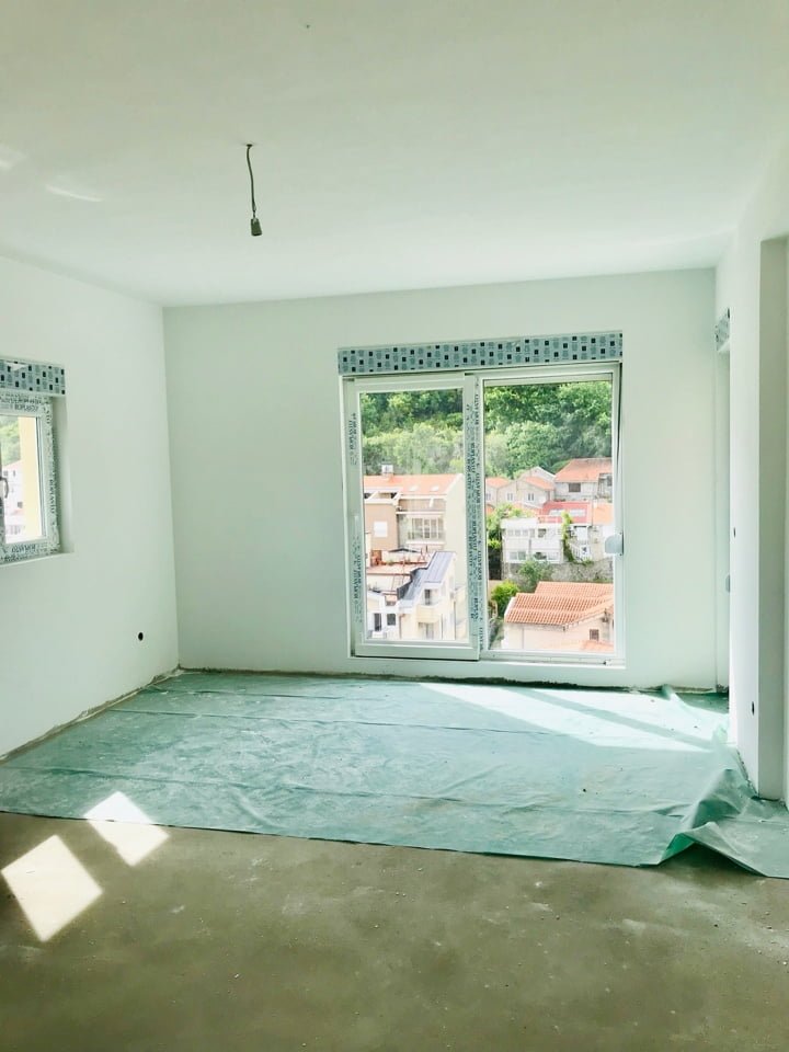 Bright unfinished living room in a new building in Bečići, Montenegro, with panoramic windows and a view of the city and mountains.