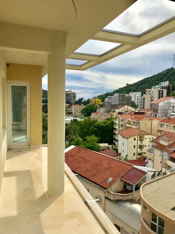 Spacious balcony with a column and a view of the town of Bečići, Montenegro, and green mountains in a residential complex.