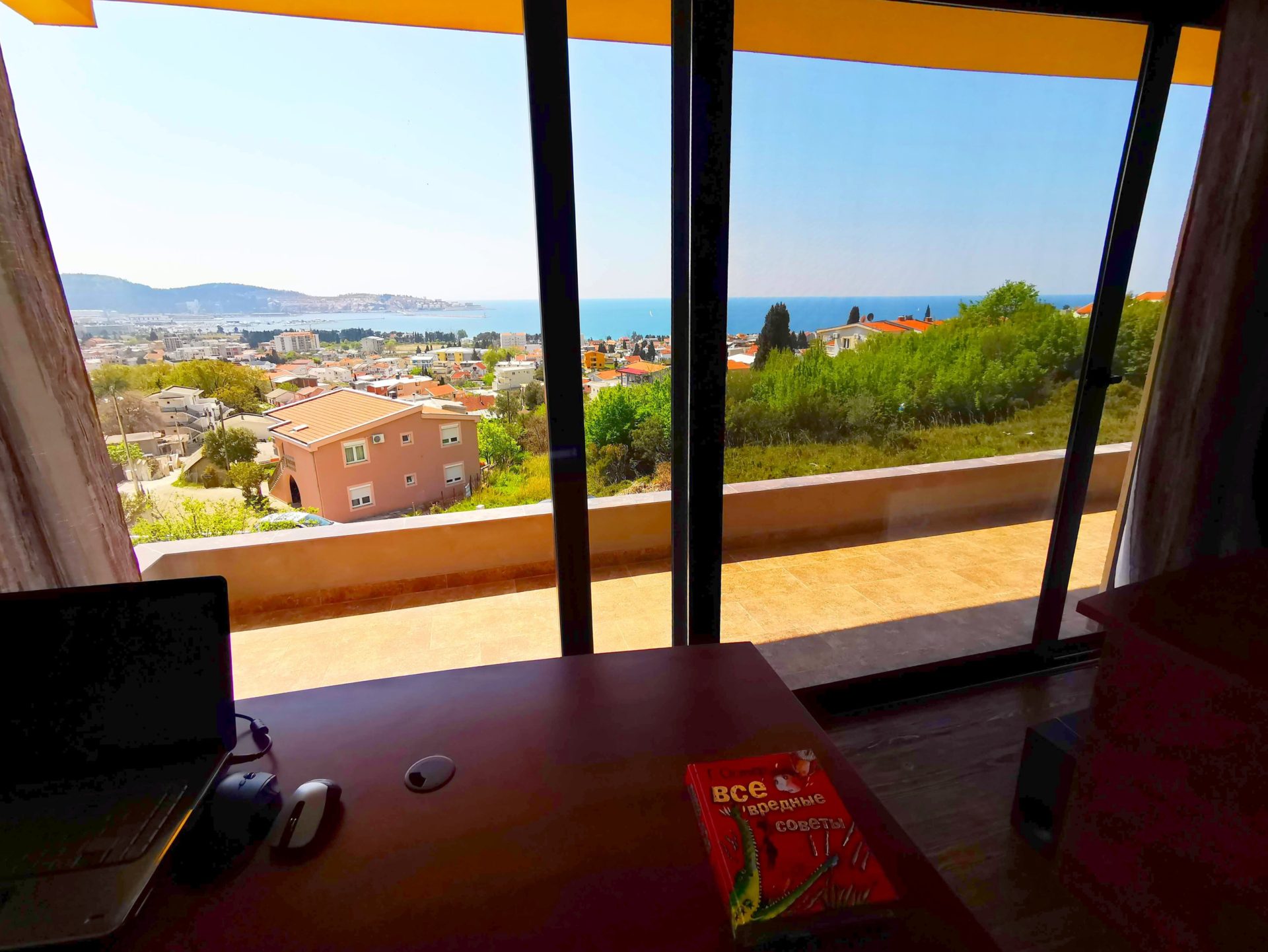 Sea and city view of Bar from the villa’s living room in Montenegro: panoramic windows, a terrace, and a workspace with a laptop.