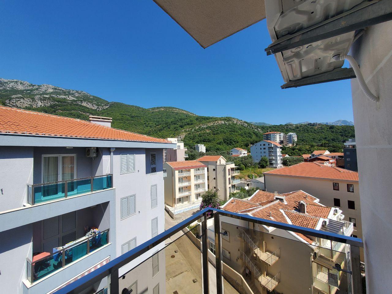 View from the balcony of a residential building in Bečići, Montenegro: a panorama of mountains, tiled roofs, and neighboring modern buildings.