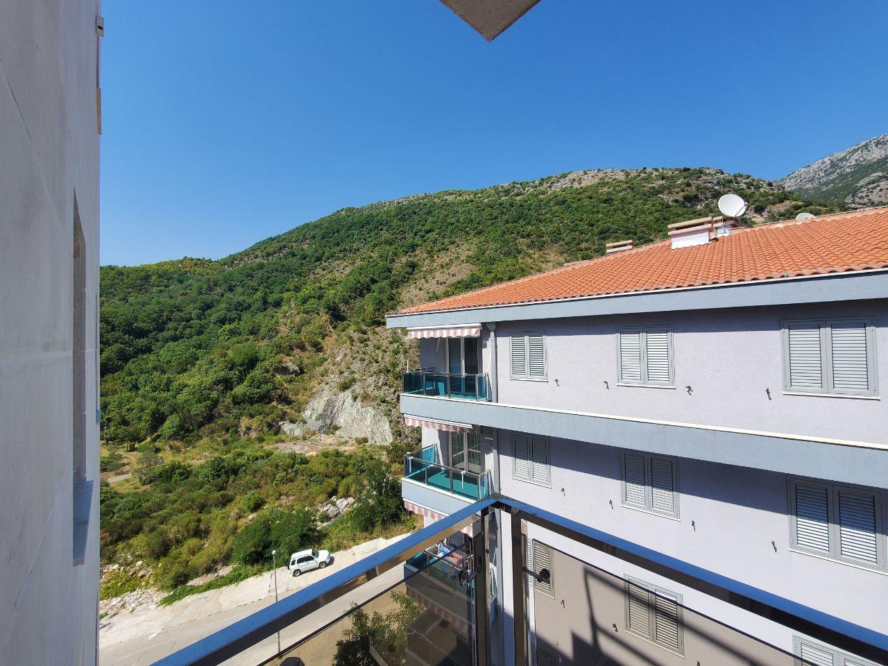 View from the balcony of a residential building in Bečići, Montenegro: a panorama of green mountains and a neighboring building with a tiled roof.