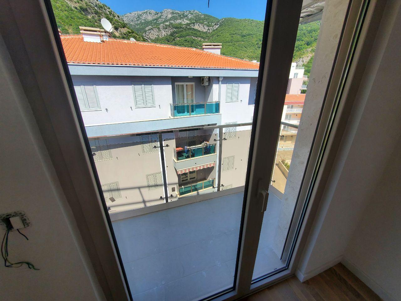 View from the balcony of an apartment in Bečići, Montenegro: panoramic windows, a modern residential building, and a mountain landscape in the background.