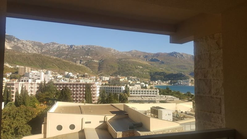 View from the balcony of an apartment in Bečići, Montenegro: a panorama of mountains, hotels, and the Adriatic Sea on a sunny day.
