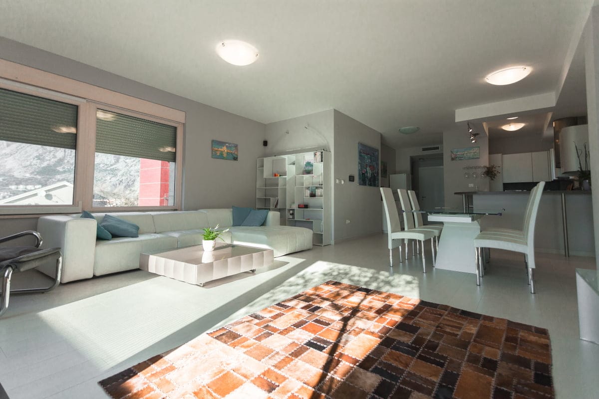 Bright living room with a white sofa and dining area in an apartment in Ljuta, Montenegro, with a mountain view from the window.