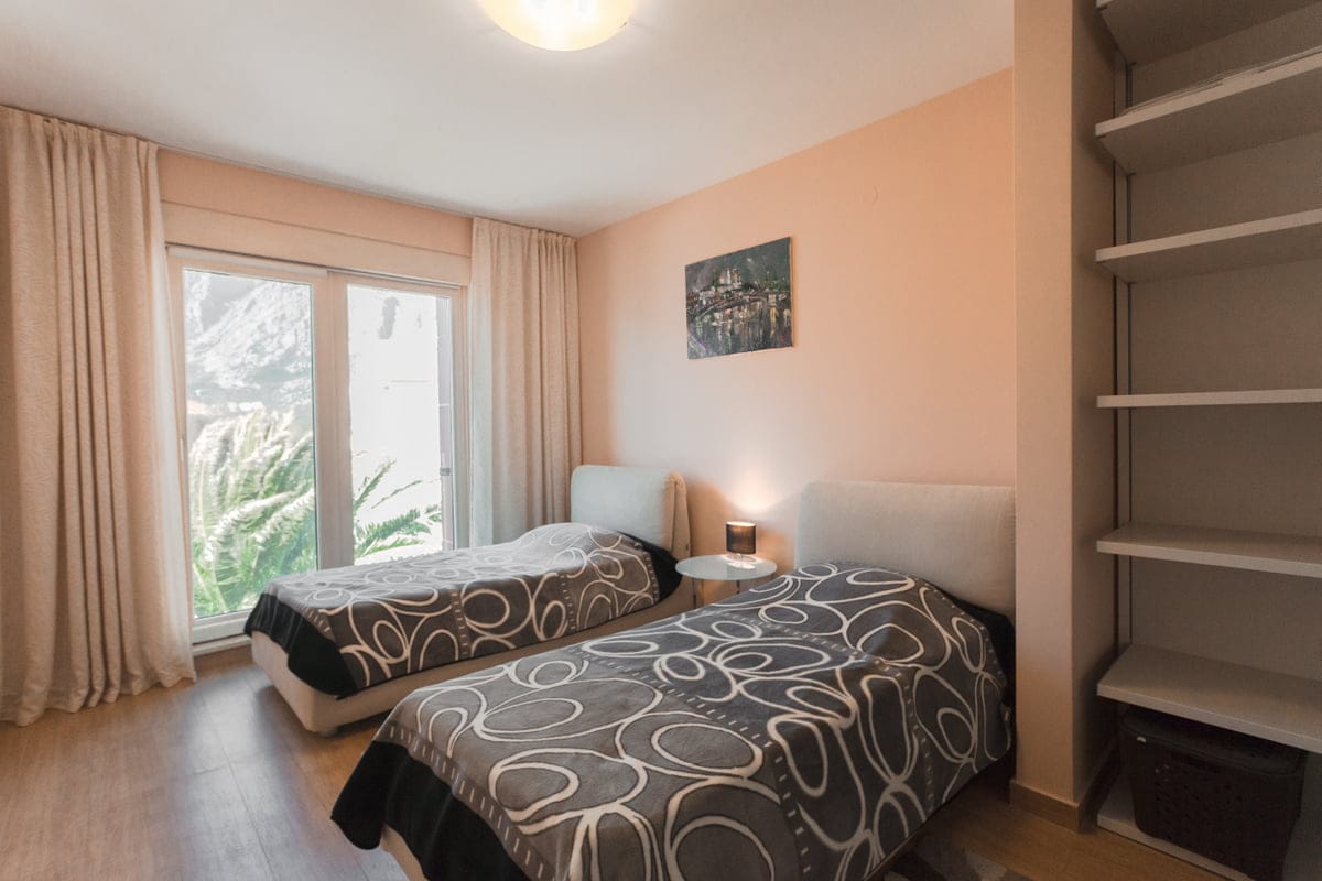Bedroom with two beds in an apartment in Ljuta, Montenegro: bright interior, panoramic window, and built-in shelves.