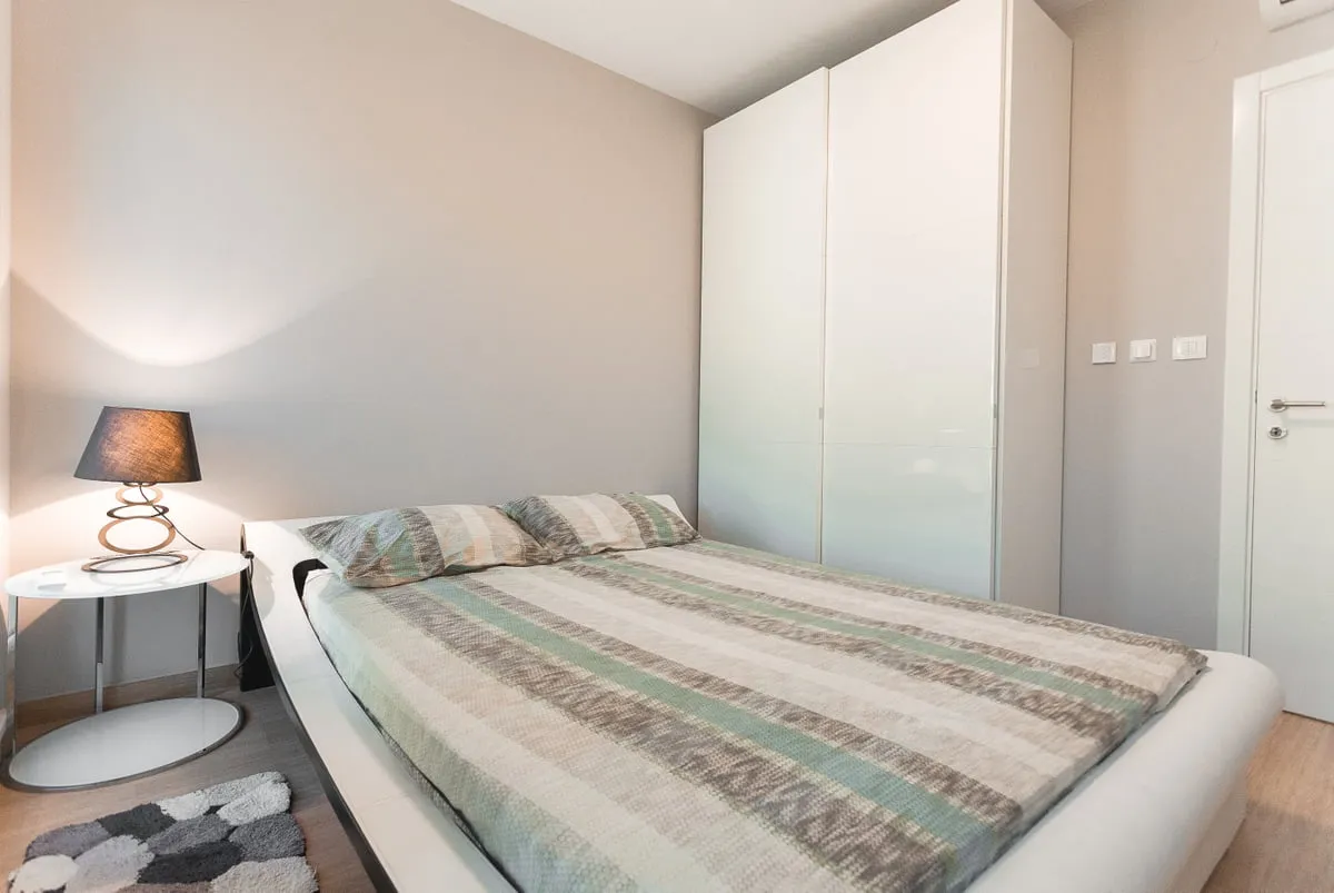Modern bedroom featuring a double bed with striped bedding, a large white wardrobe, and a side table with a spiral lamp.
