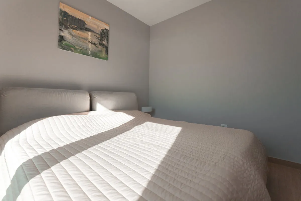 Bedroom featuring a double bed with a light quilted cover and an abstract painting on a grey wall.