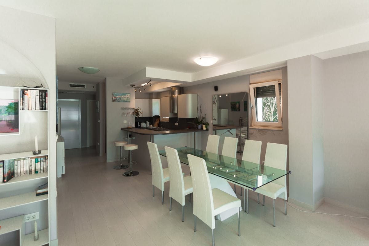 Modern living room with dining area and kitchen in an apartment, Ljuta, Montenegro: glass table, white chairs, and a breakfast bar.