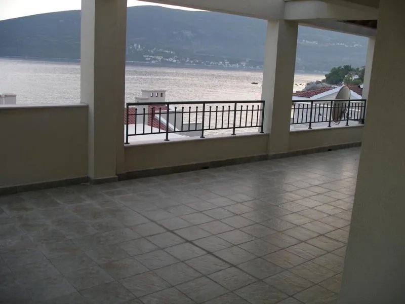 Tiled terrace with black metal railing overlooking the sea and mountains in the distance.