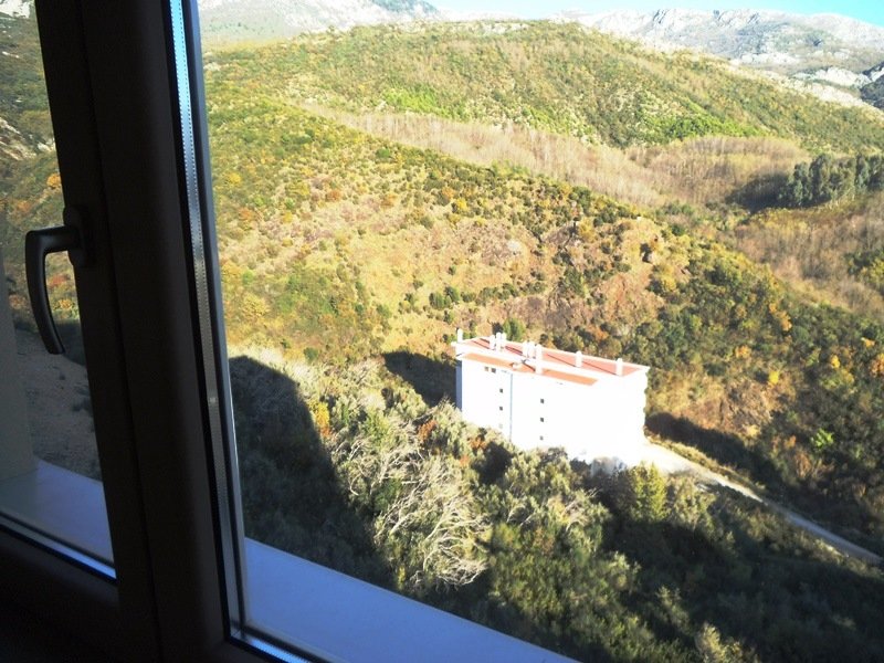 View from the apartment window in Bečići, Montenegro: a panorama of green mountains and a neighboring residential building with a red roof.