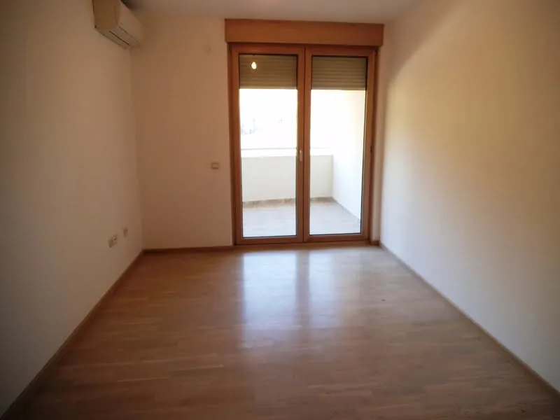 Empty room with laminate flooring and glass door to balcony. White walls, AC unit visible, no furniture present.
