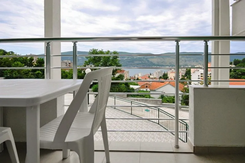 Balcony with white plastic chairs and table, metal railing, overlooking a town and lake.