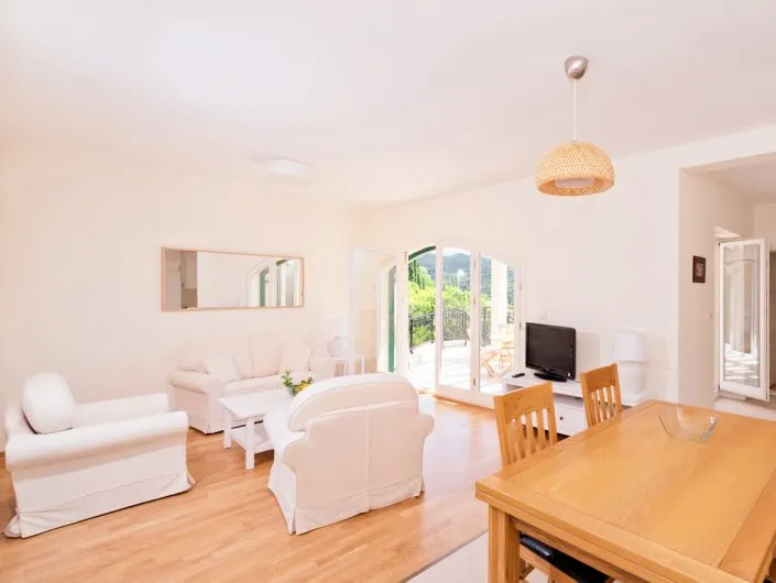 Bright room with white sofas, wooden table, TV, and glass doors leading to a balcony.