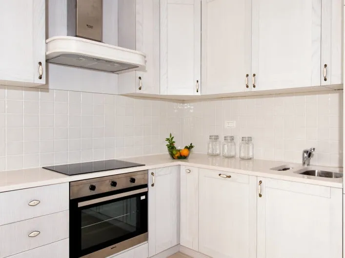 White kitchen with built-in oven, cooktop, hood, and sink on a light countertop with upper cabinets.