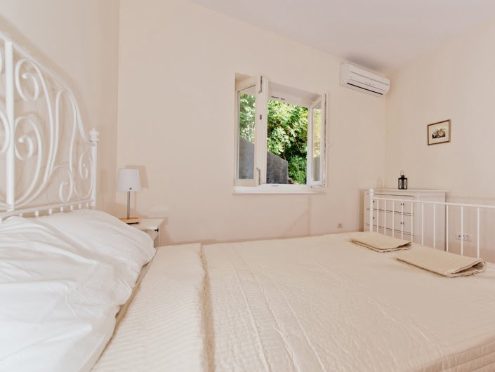 Bright bedroom with a white wrought-iron bed in an apartment in Herceg Novi, Montenegro, with a view of the green garden from the window.