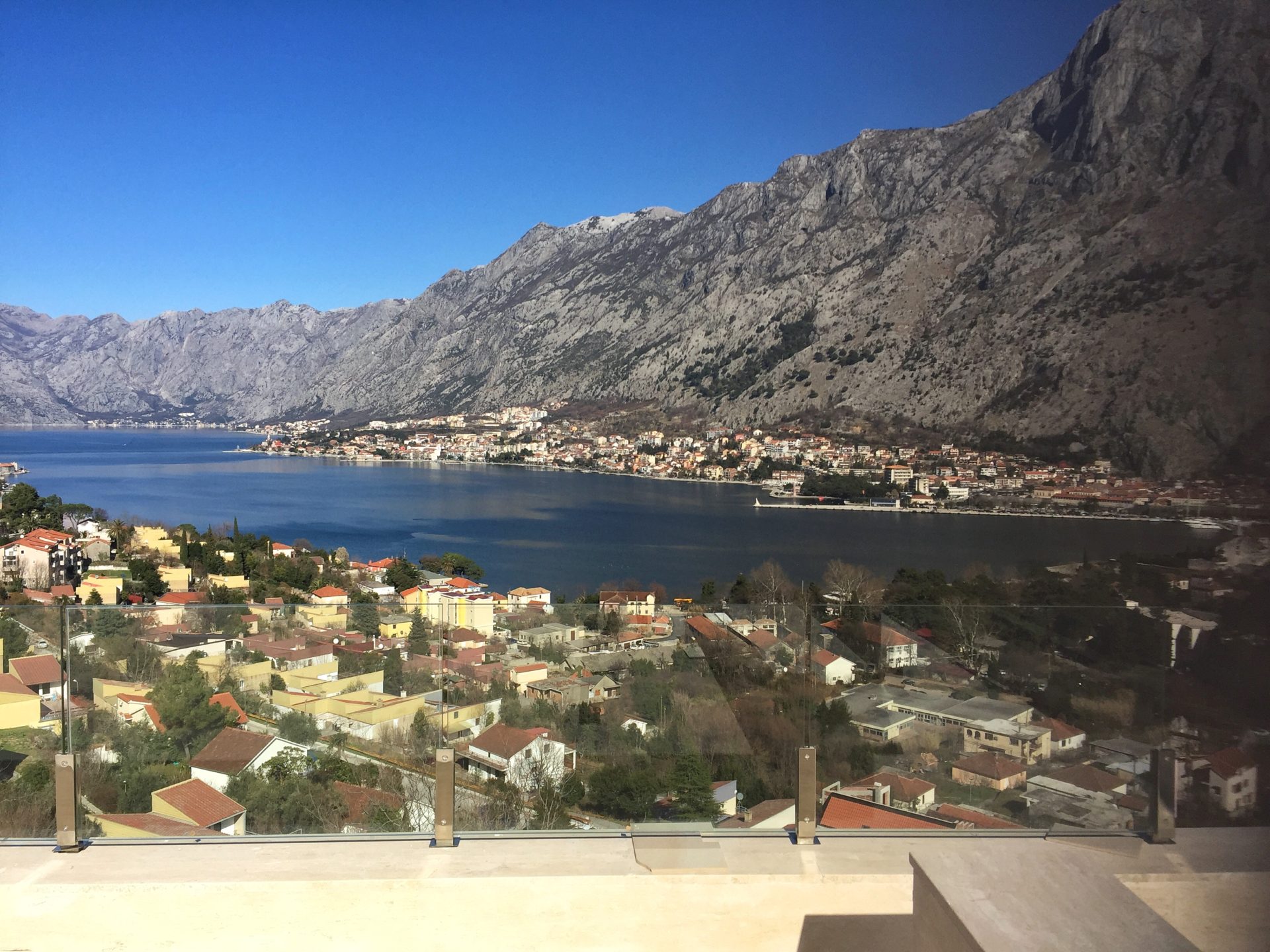 Panoramic view of the Bay of Kotor and the mountains from the terrace of a residential complex in Kotor, Montenegro, with a glass railing.