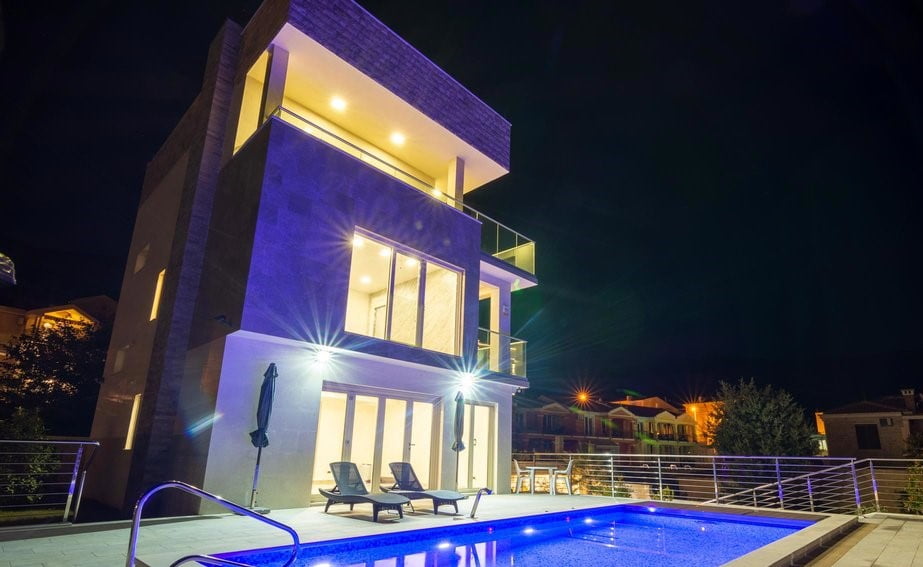 Modern three-story villa with a swimming pool and night lighting in Bečići, Montenegro. Facade view with a terrace.