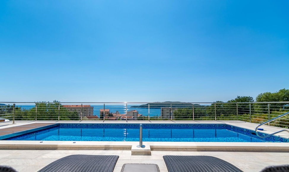 Outdoor swimming pool on a villa terrace in Becici, Montenegro, with a panoramic view of the Adriatic Sea and mountains.