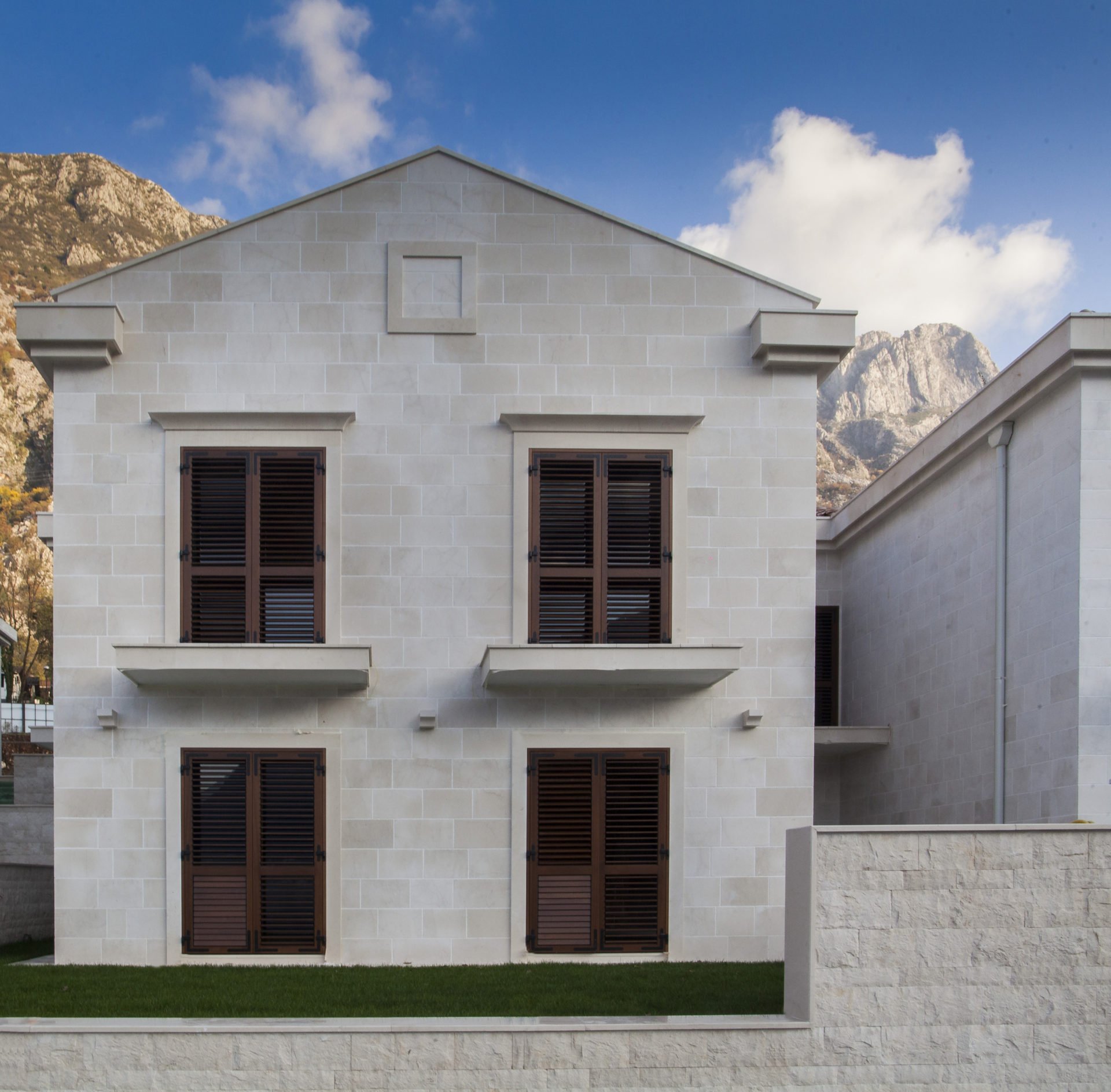 Facade of a modern villa made of light stone with brown shutters in Dobrota, Montenegro, against a backdrop of mountain scenery.