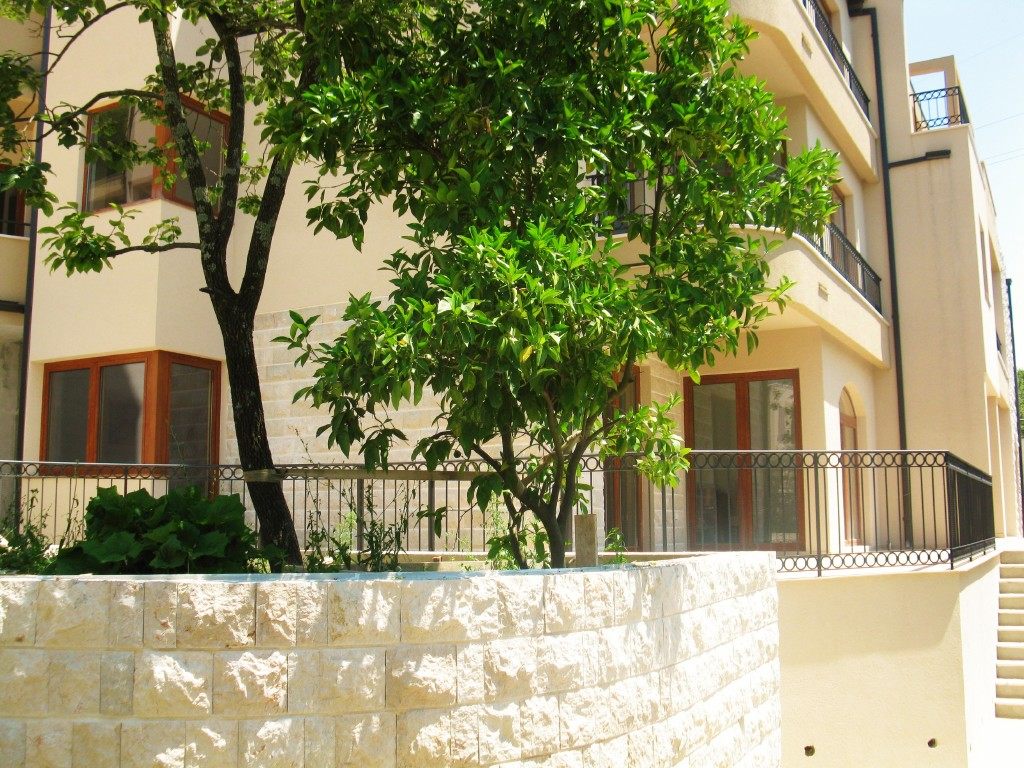 Facade of a residential complex in Igalo, Montenegro: stone cladding, terraces with wrought-iron railings, and a green tree in the courtyard.