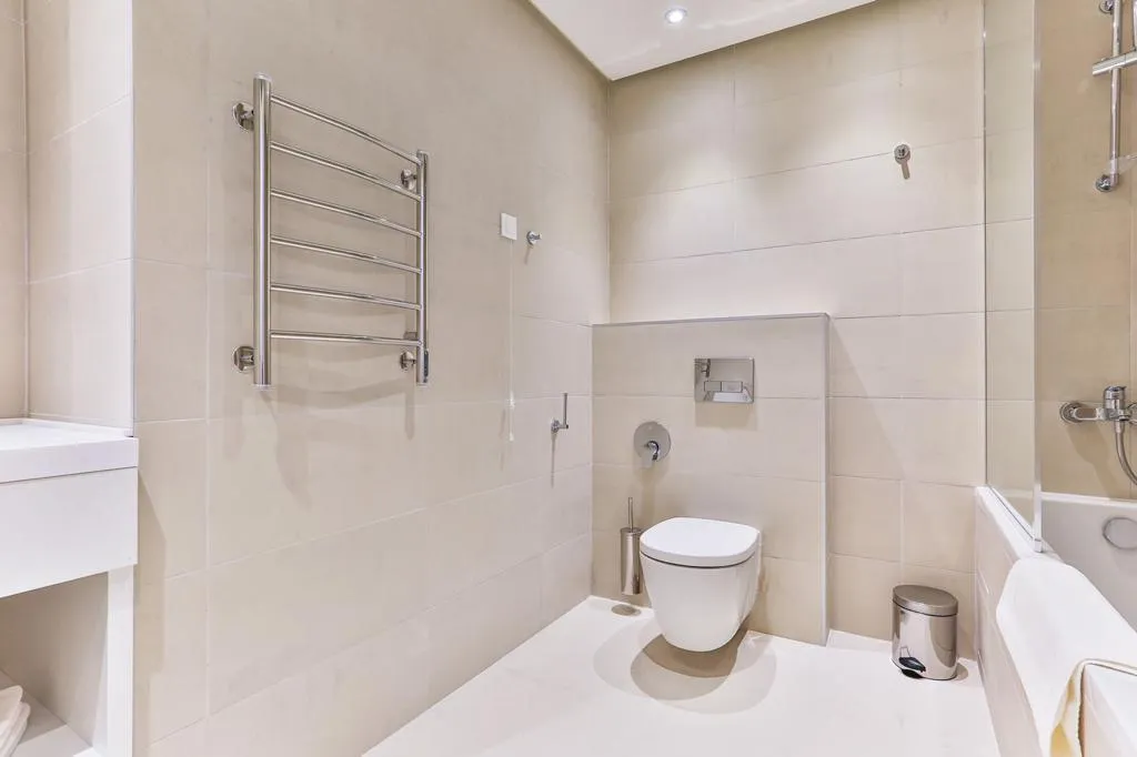 Modern bathroom featuring a wall-mounted toilet, heated towel rail, and bathtub against beige tiles.