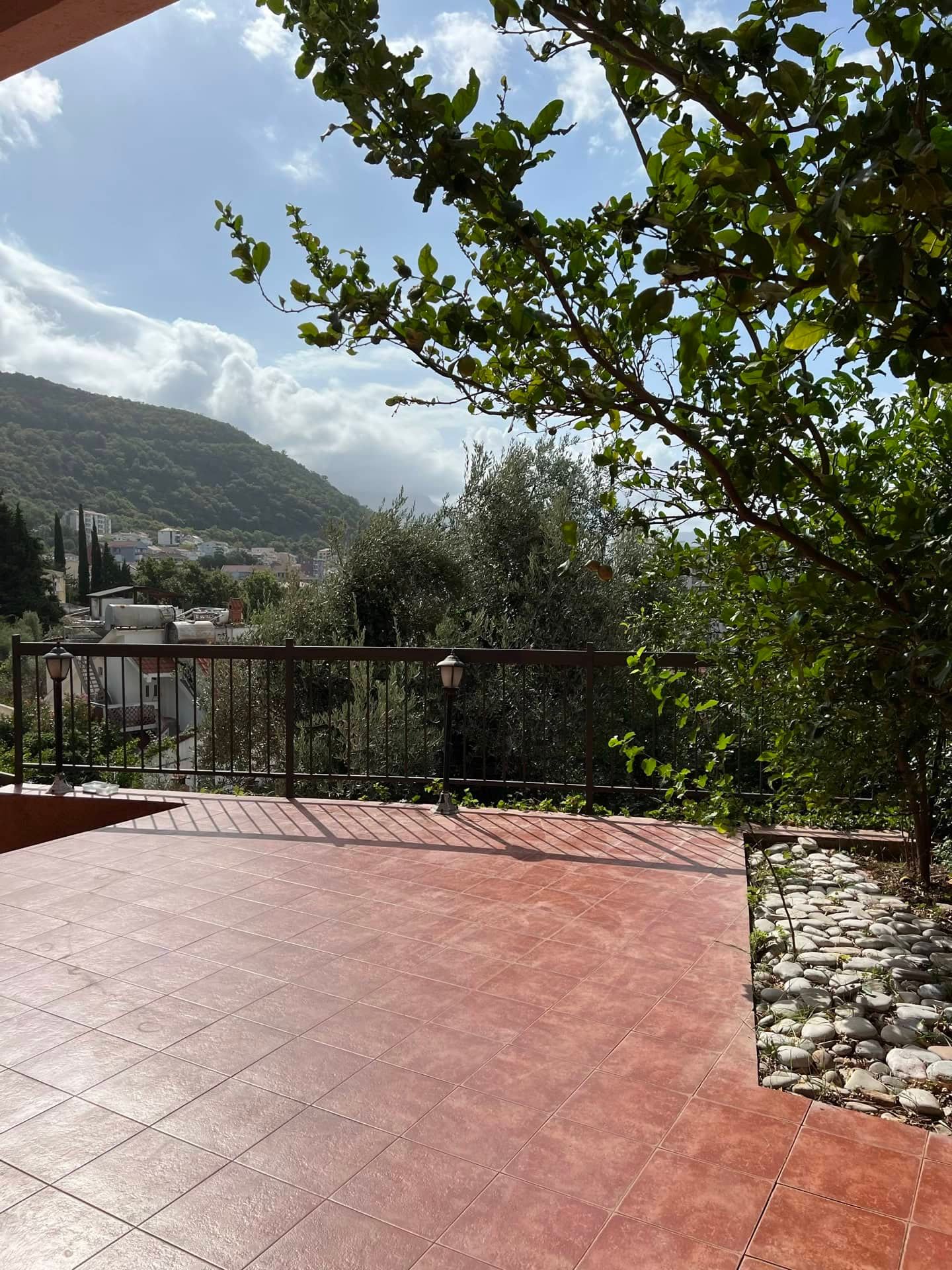 Spacious tiled terrace with mountain views in an apartment in Petrovac, Montenegro, with green trees and a railing.