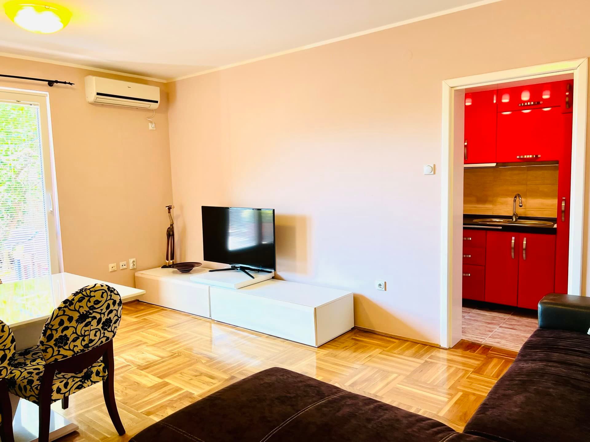 Living room with parquet flooring, TV area, and a bright red kitchen in an apartment in Petrovac, Montenegro.