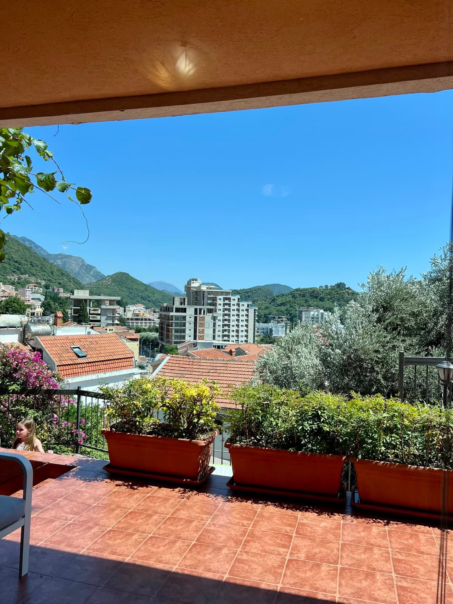 Tiled terrace with large planters and a view over city rooftops and green mountains under a blue sky.
