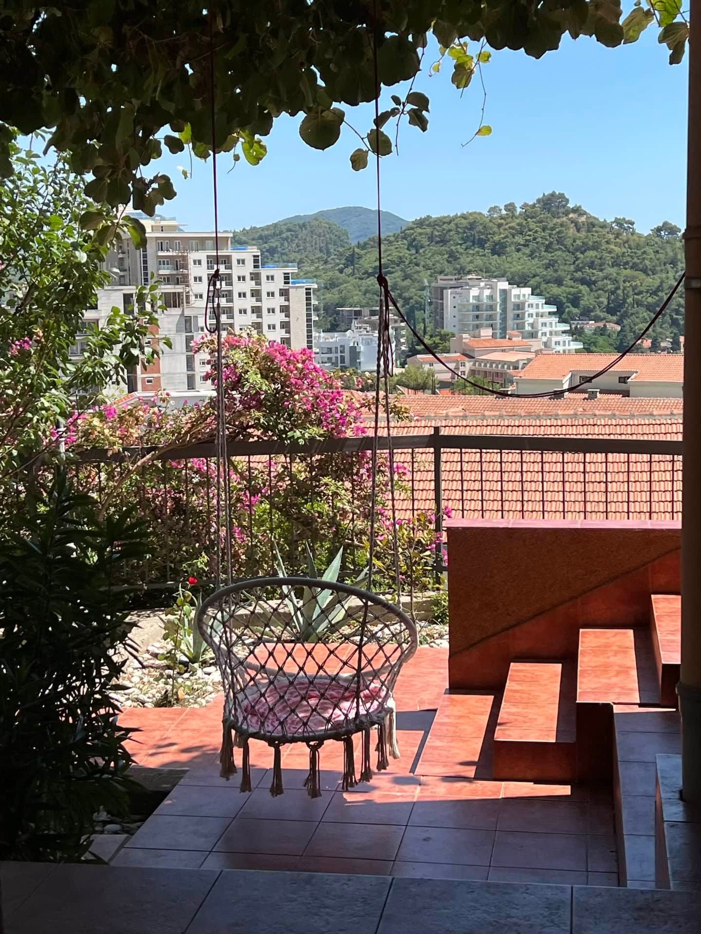 Duplex terrace in Petrovac, Montenegro: hanging chair, mountain, sea and city views among flowering plants.
