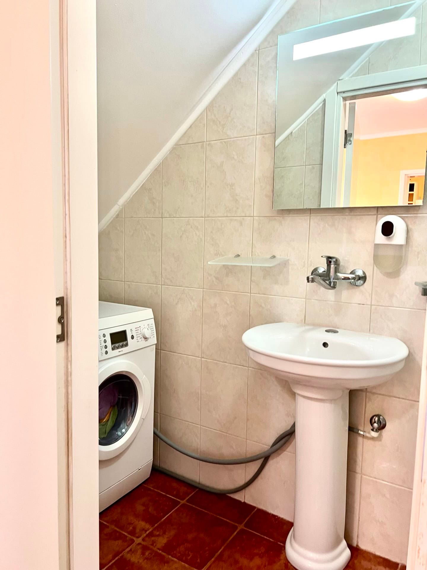 Bathroom in a duplex in Petrovac, Montenegro: pedestal sink, mirror, and washing machine under the sloped ceiling.