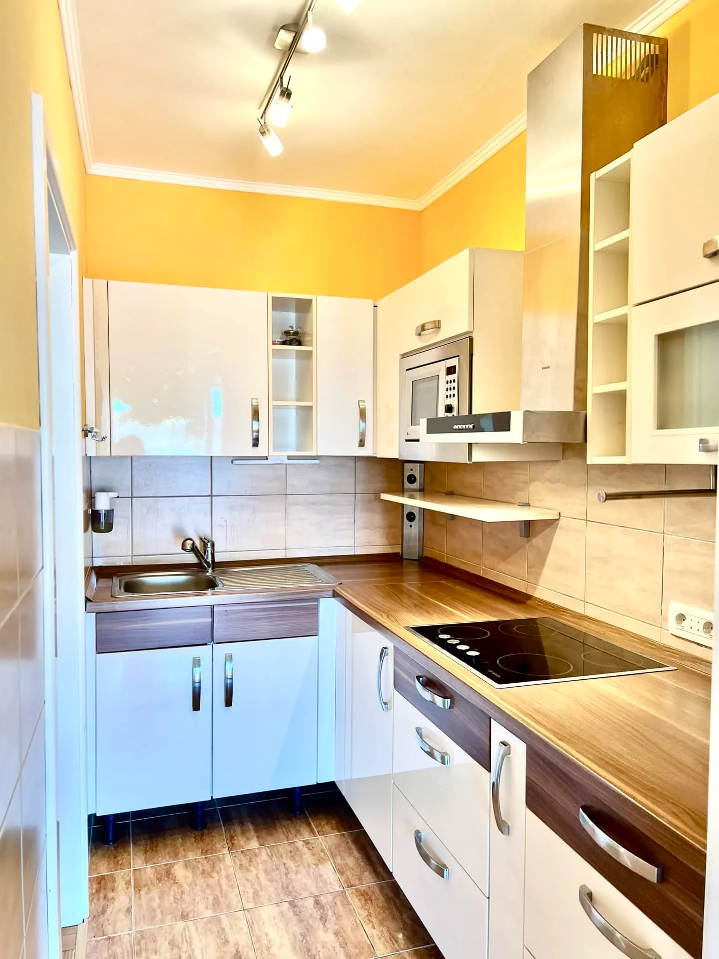 Corner kitchen with white cabinets, wood countertop, sink, induction cooktop, microwave, and tiled walls.