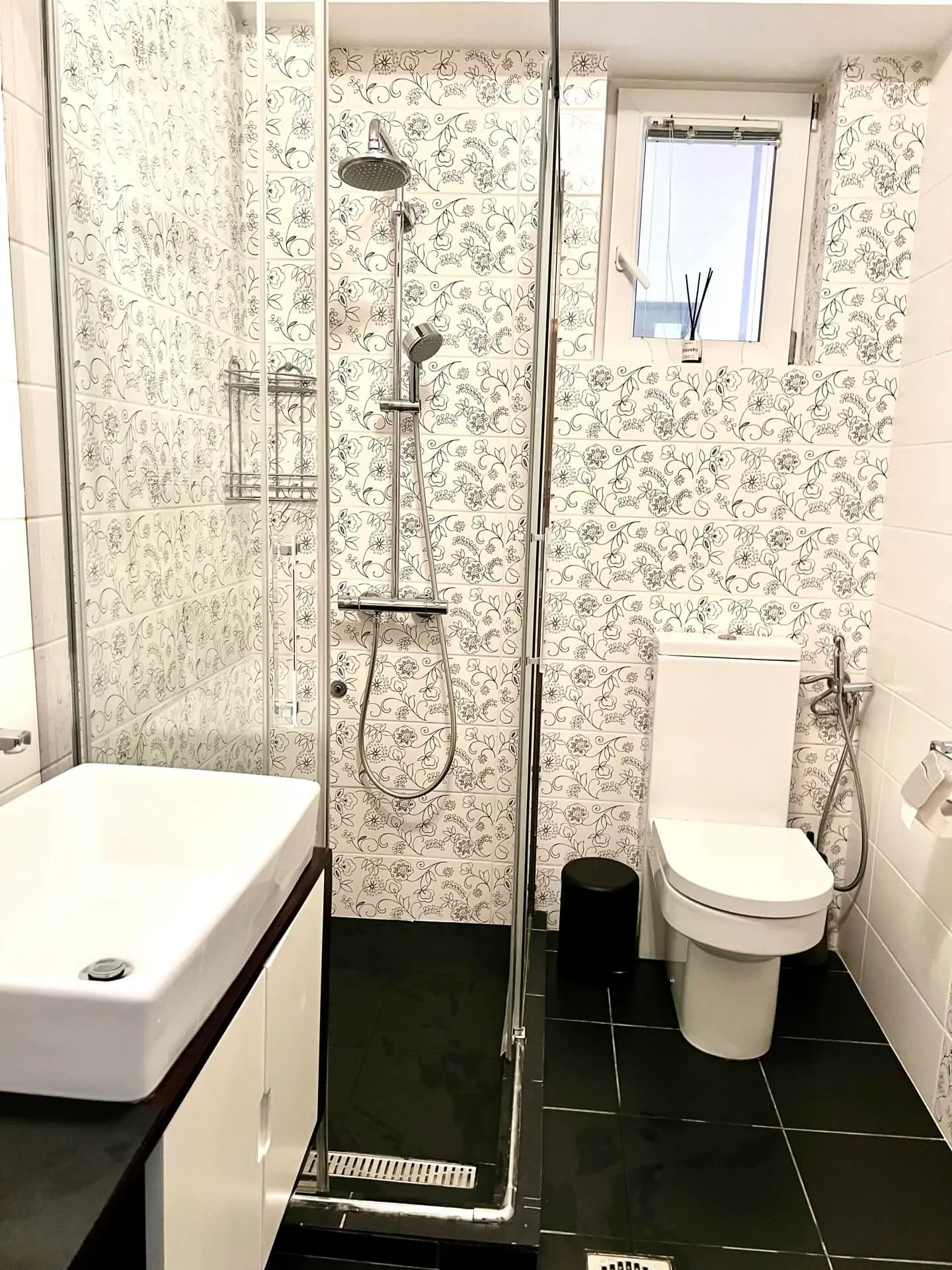Bathroom with shower, toilet, and sink featuring floral wallpaper and black floor tiles.