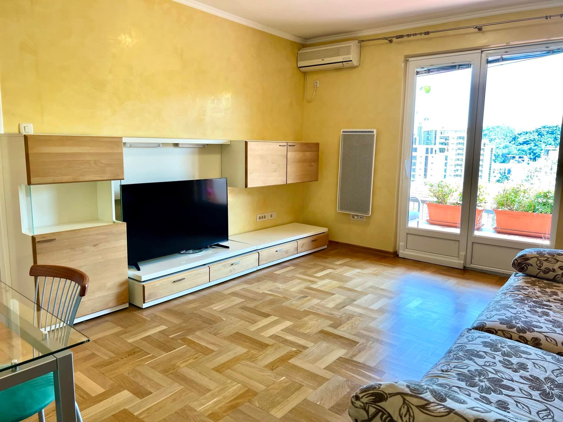 Living room in an apartment in Petrovac, Montenegro: parquet flooring, a modern TV wall unit, and access to a terrace with city views.