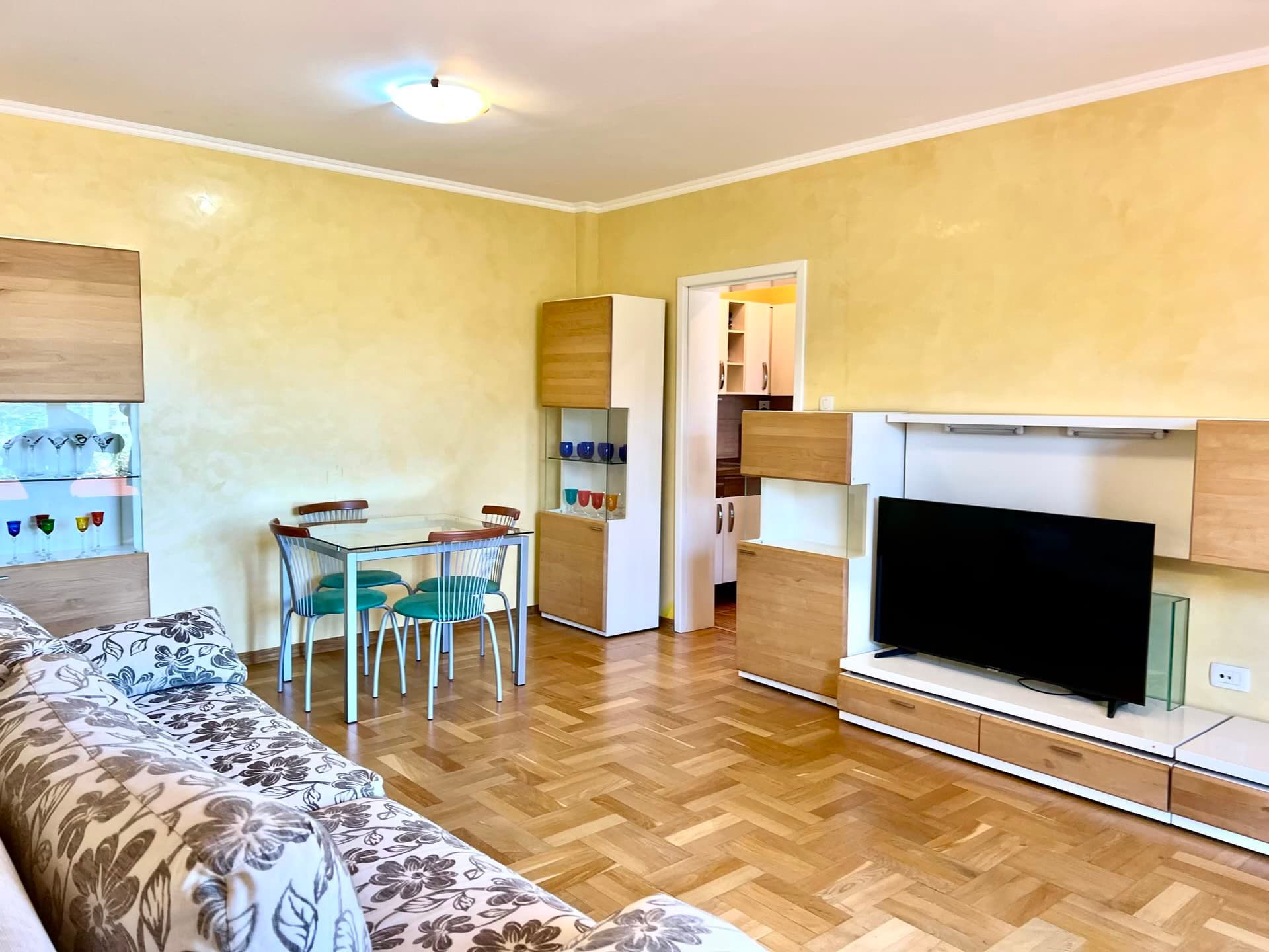 Living room with parquet flooring and dining area in an apartment in Petrovac, Montenegro: a soft sofa, TV unit, and light finishes.