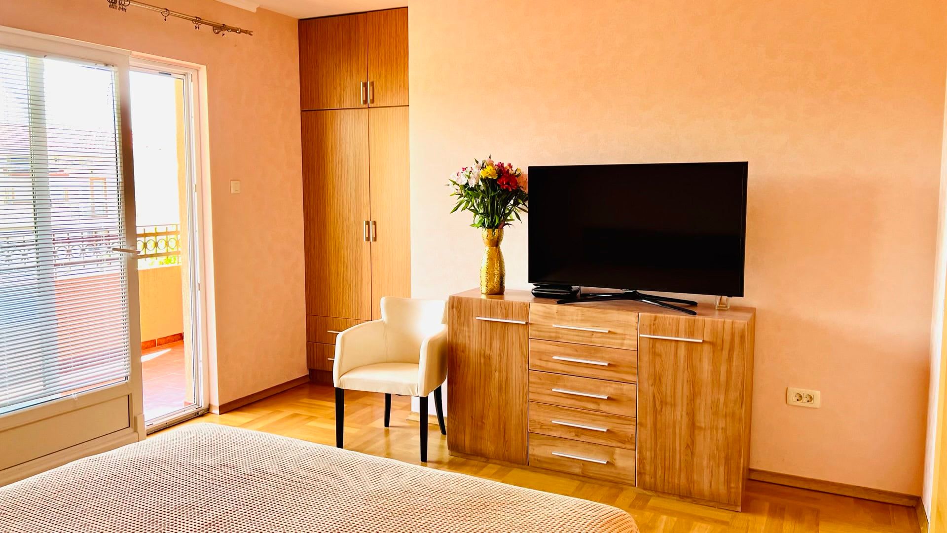 Cozy bedroom in an apartment in Petrovac, Montenegro: wooden furniture, TV, armchair, and access to a balcony with a view.