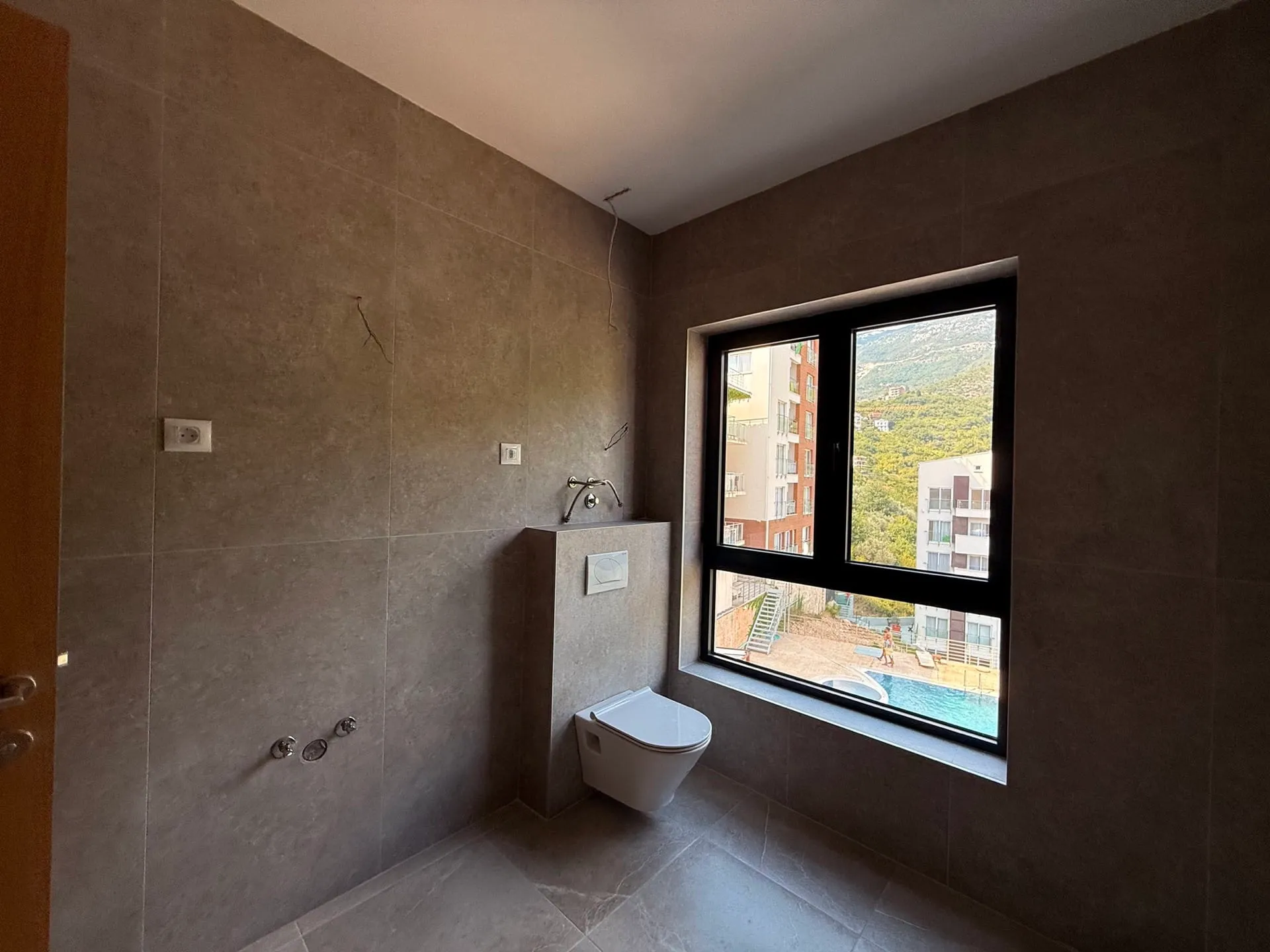 Bathroom with grey tiles, wall-mounted toilet, and a window overlooking a pool and hills.