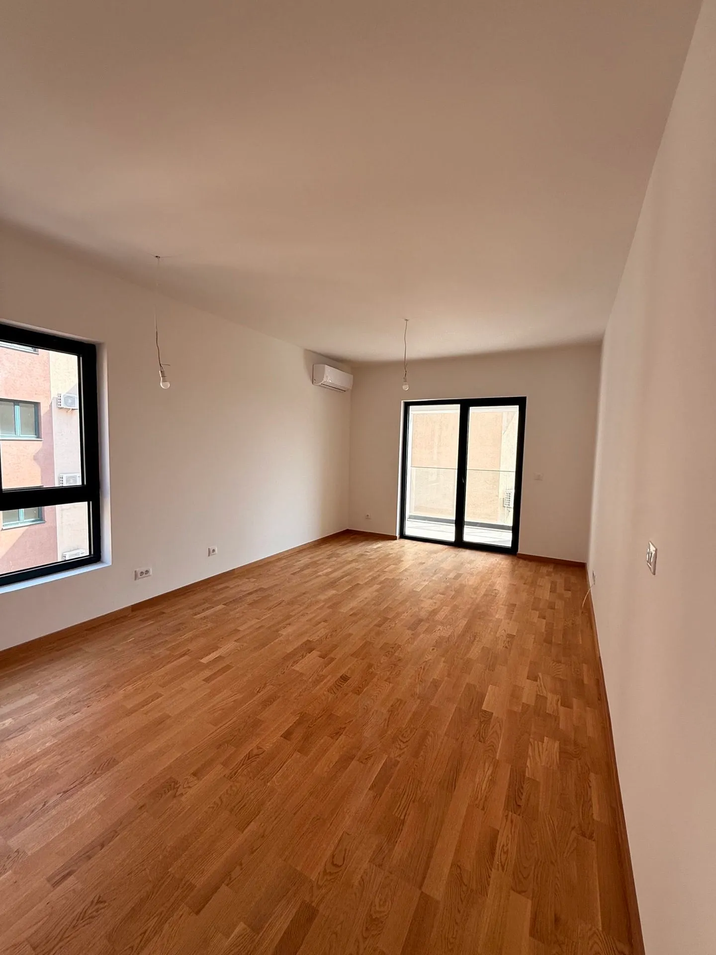 Empty room with wooden flooring, a window, and a glass door leading to a balcony. Hanging bulbs and AC unit visible.