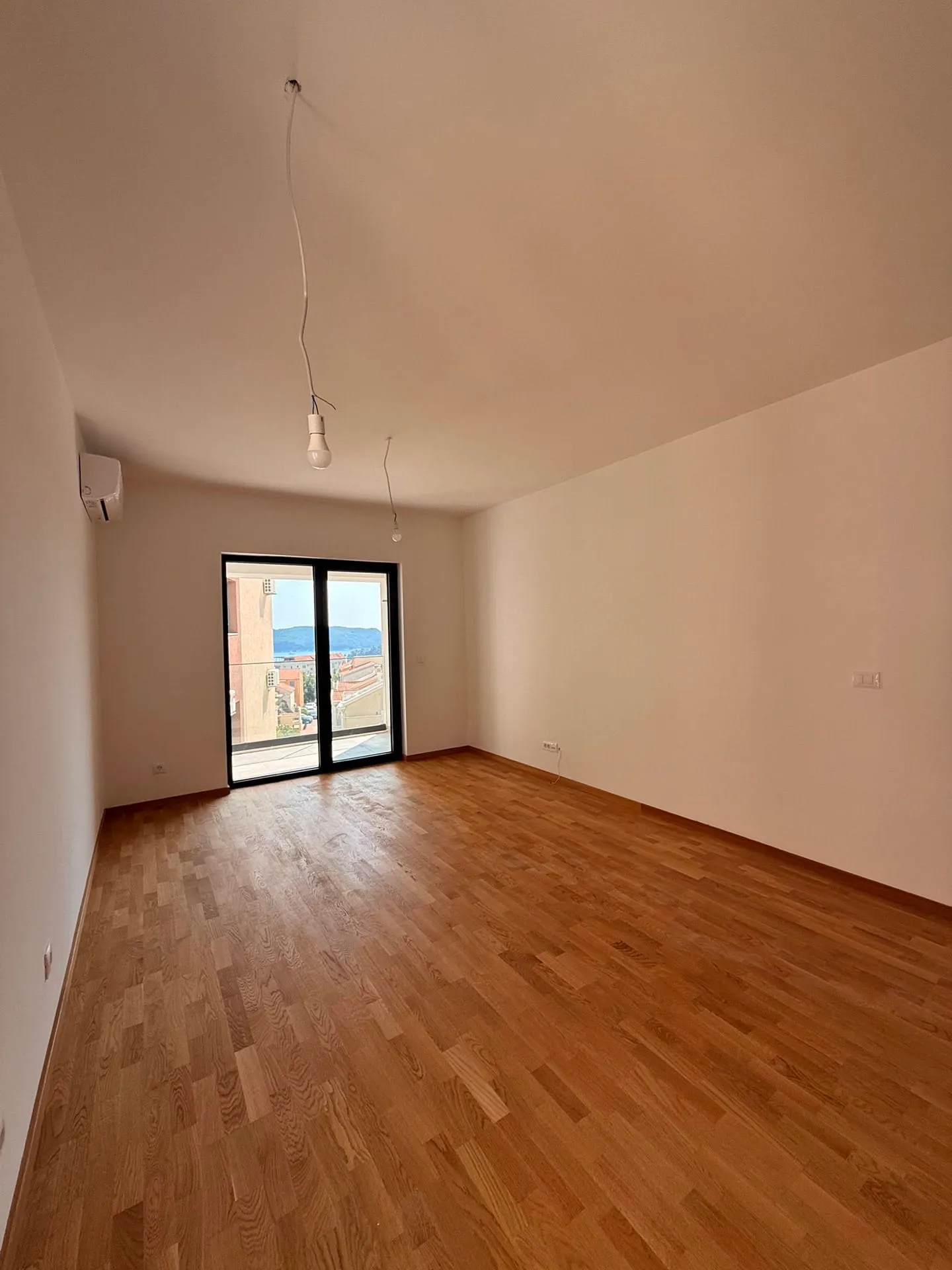 Empty room with wooden flooring, white walls, air conditioner, and sliding glass door to balcony.