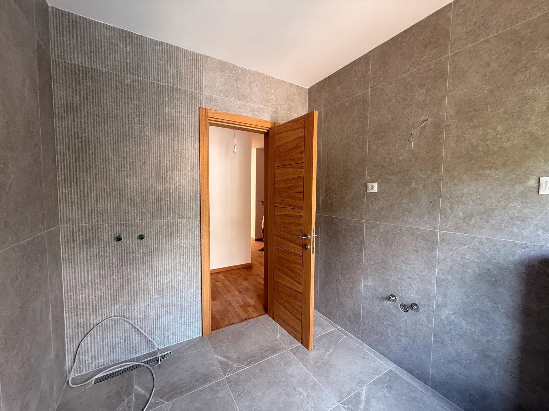 Unfinished bathroom with gray wall tiles, exposed water pipes, a floor drain, and a wooden door.
