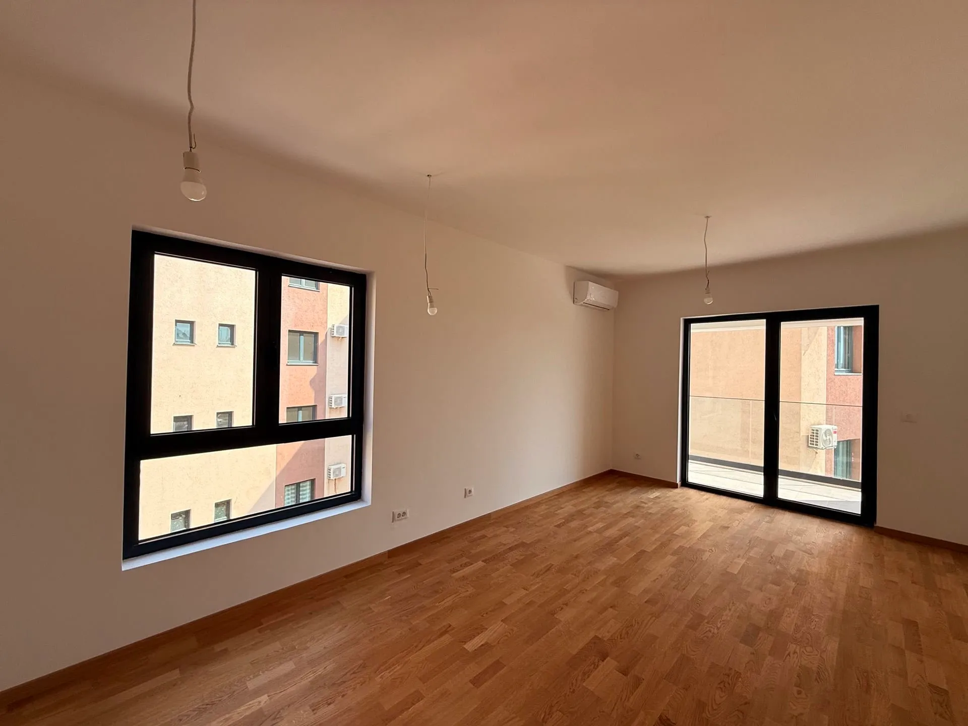 Empty room with wooden flooring, a large window, and a sliding door leading to a balcony. Features an AC unit and hanging bulbs.