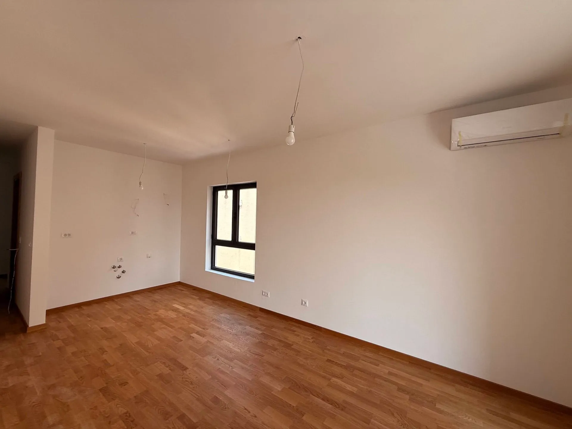 Empty room with wood flooring, white walls, a window, and an AC unit. Hanging light bulbs are visible.