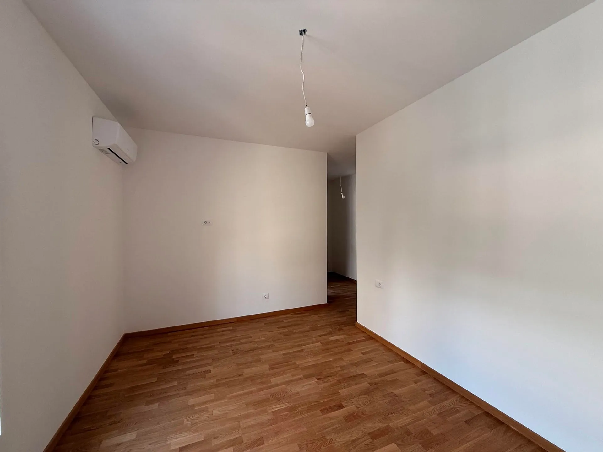 Empty room with laminate flooring, white walls, a hanging bare bulb, and a wall-mounted air conditioner.