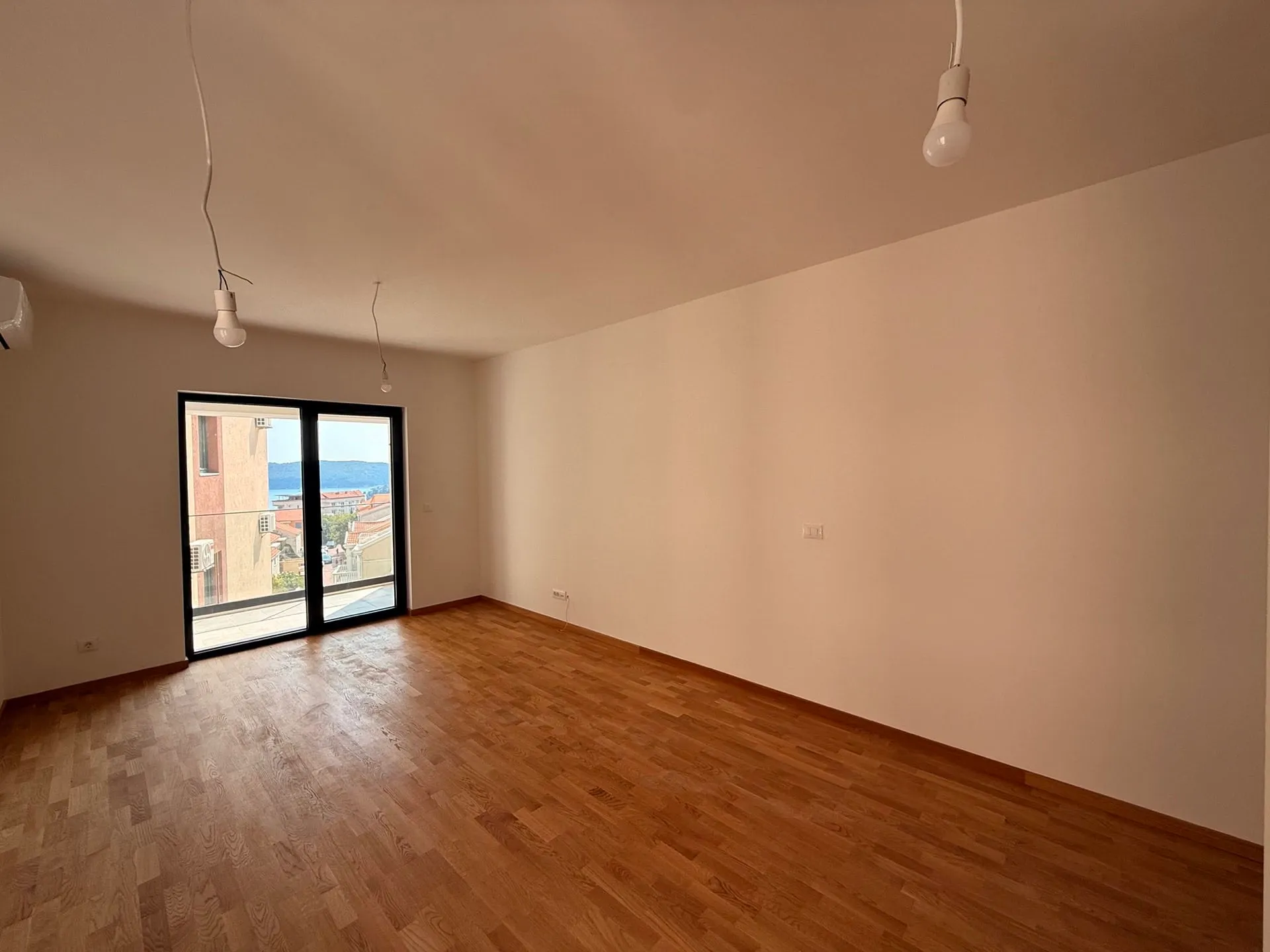 Empty room with wooden flooring and white walls. Hanging bulbs visible and glass door leading to a balcony.