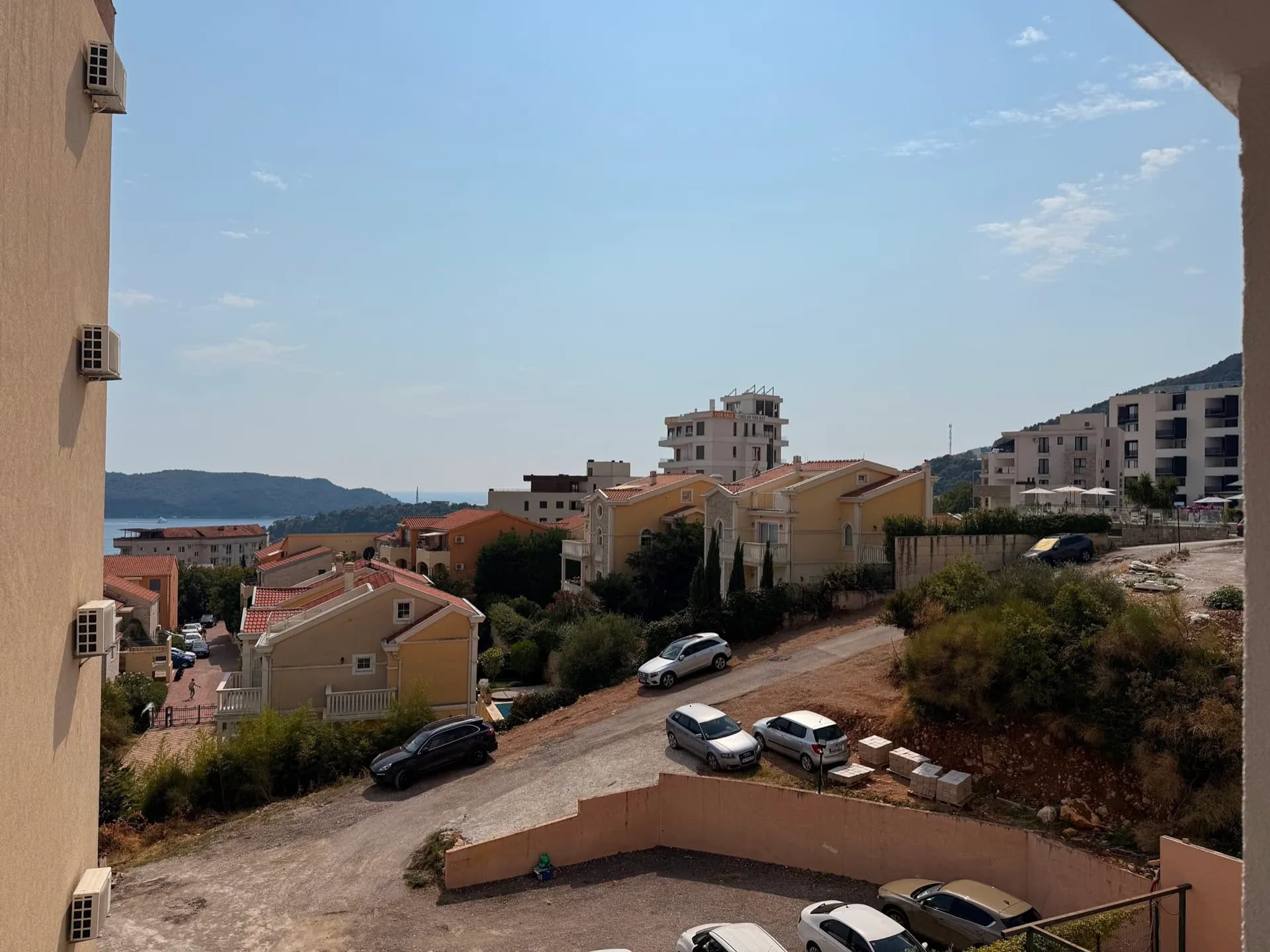 View from a balcony showing residential houses, parked cars on a dirt road, and the sea in the distance.