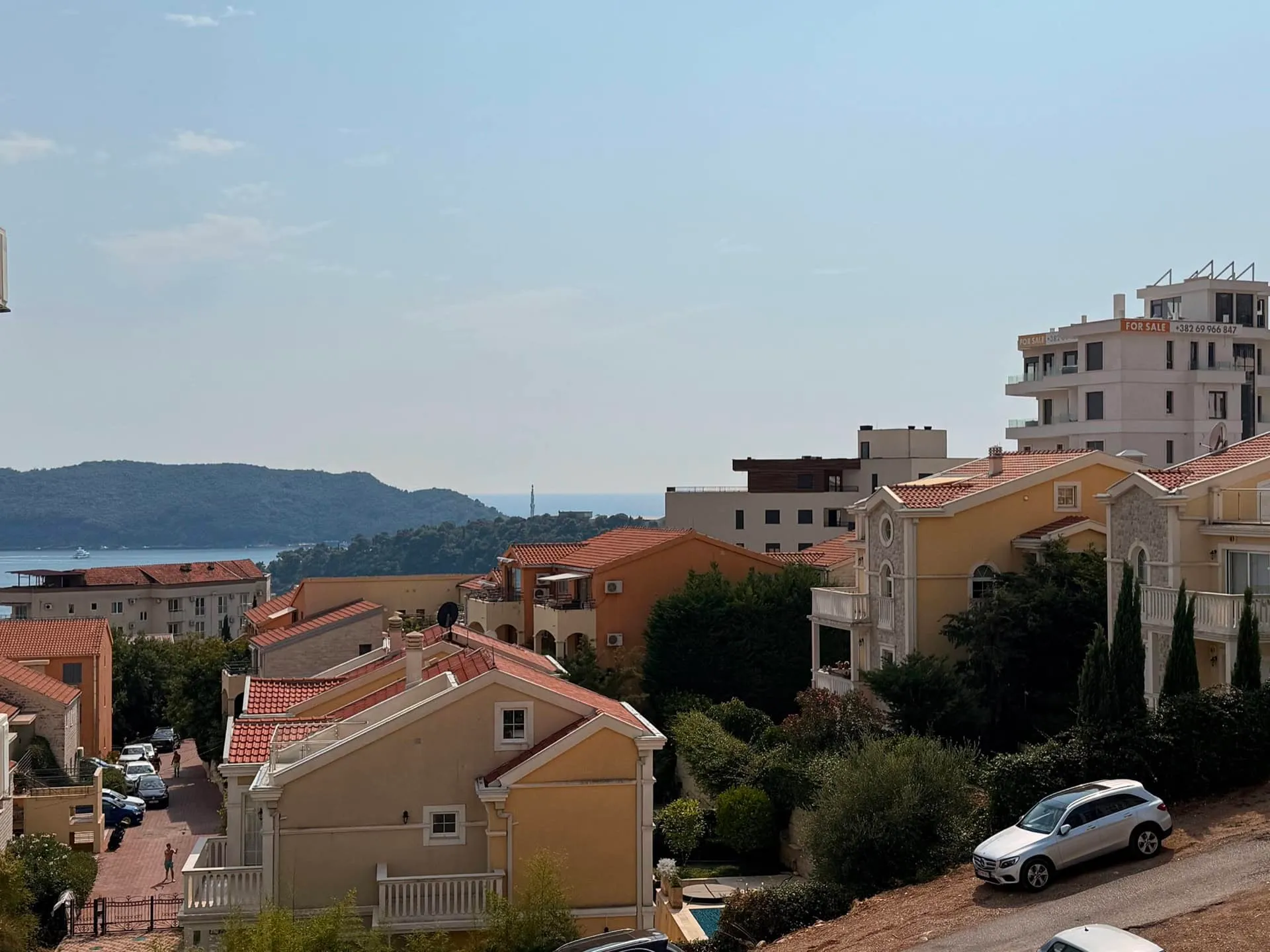 Panoramic view of residential rooftops, a street with cars, and a sea landscape with hills.