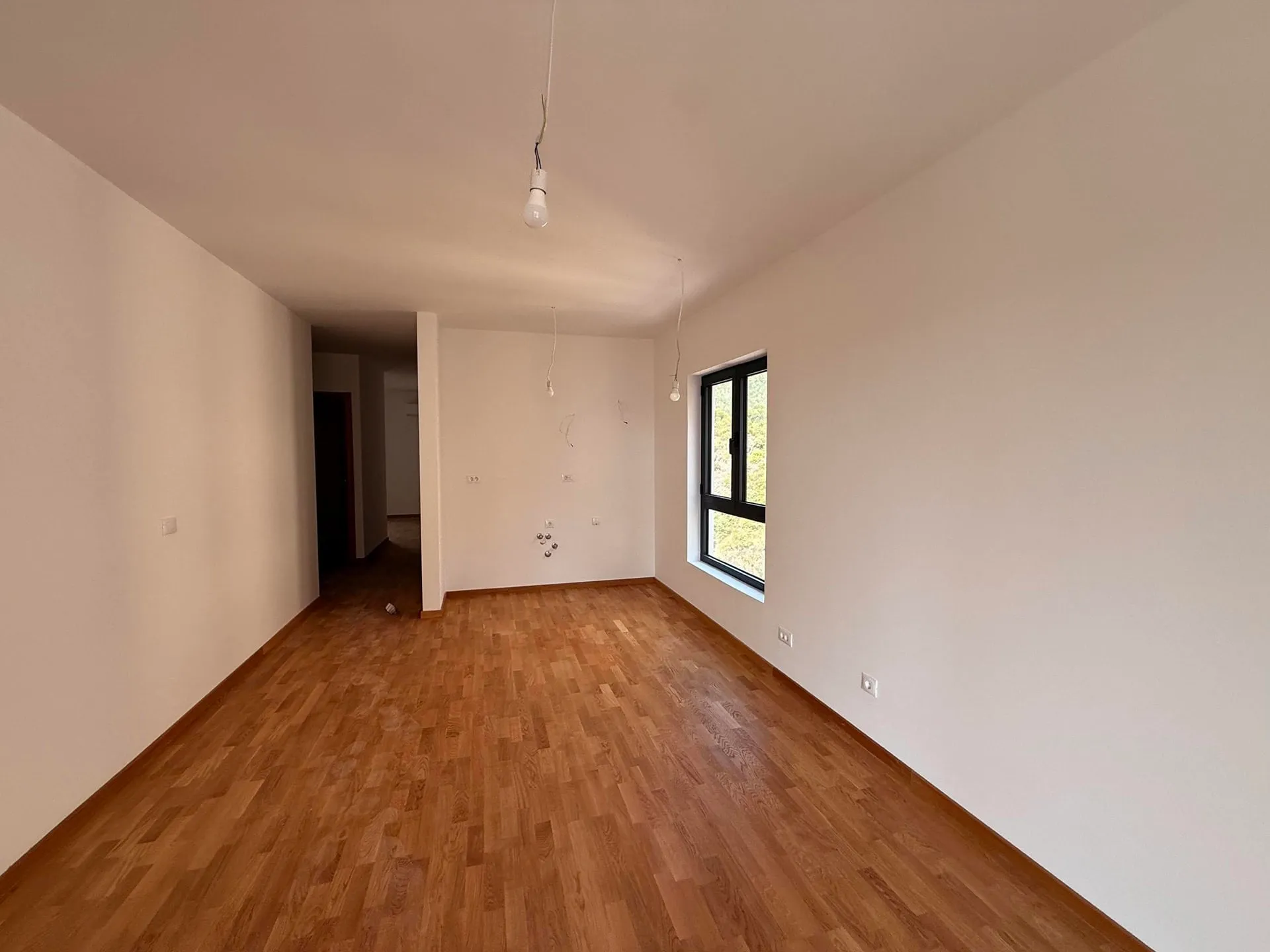 Empty room with wood laminate flooring, white walls, a window, and bare hanging light bulbs.