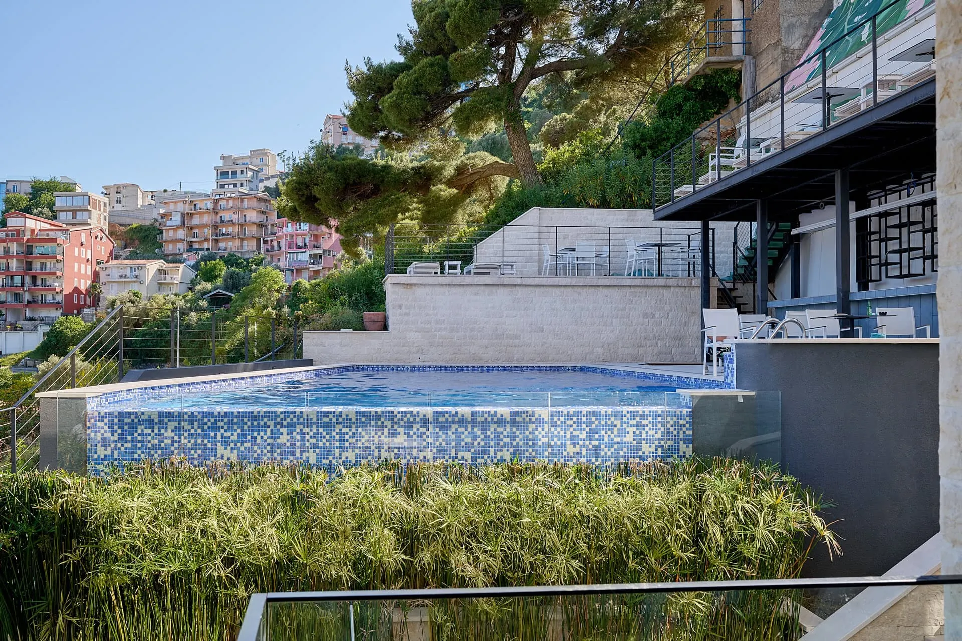 Outdoor pool with blue mosaic tiles and glass barrier, surrounded by tall grass and a terrace with white furniture.